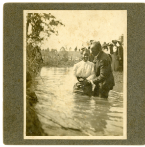 Sepia tone photograph of a man and woman standing in a river, with the water up to their thighs. They are preparing to lean the woman back into the water.