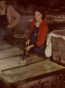 Color photograph of a woman standing at a specialty table and holding a shovel like tool. To her left, a thick liquid is pouring out of a barrel on to the table.
