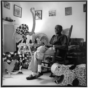Black and white photograph of a man seated in a rocking chair. He is surrounded by wooden sculptures.