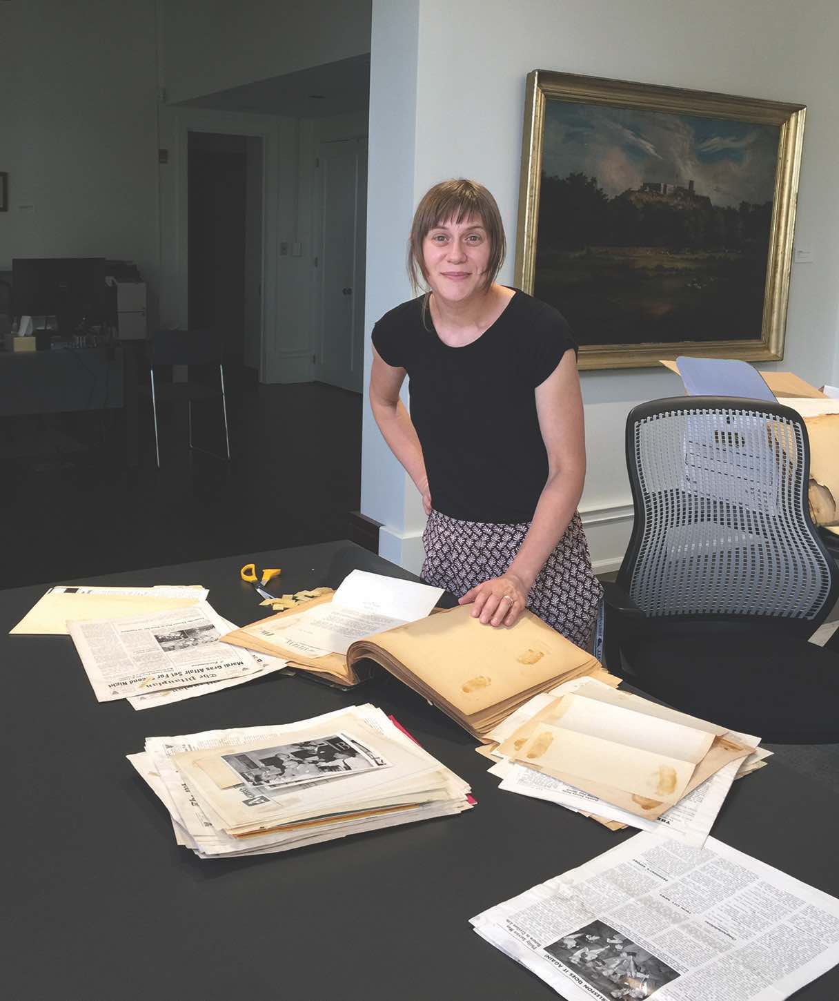 Dr. Abigail Glogower wears a black shirt and gray pants. She is standing by a black table with old documents on it. There is a landscape painting in the background.