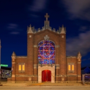 A small brick church with a large, colorful stained glass window and a red door is illuminated at night, creating a warm and inviting glow.