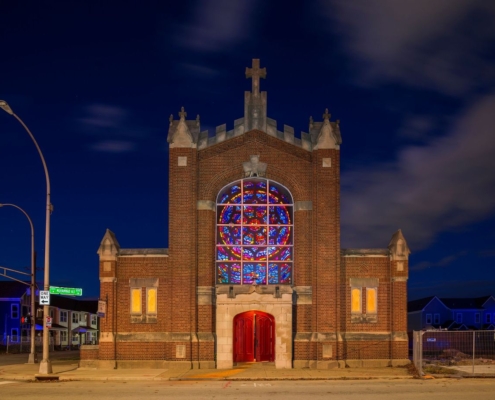 A small brick church with a large, colorful stained glass window and a red door is illuminated at night, creating a warm and inviting glow.