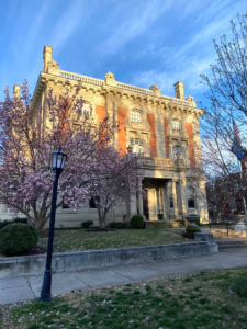 An image of the historic Ferguson Mansion with a flowering magnolia tree in the foreground. The house is red brick and white stonework. The sky is blue in the background.