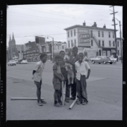 Five smiling children stand on a city street holding a dog. Background features vintage cars and storefronts, creating a nostalgic urban scene.