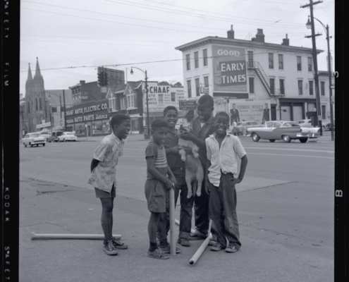 Five smiling children stand on a city street holding a dog. Background features vintage cars and storefronts, creating a nostalgic urban scene.