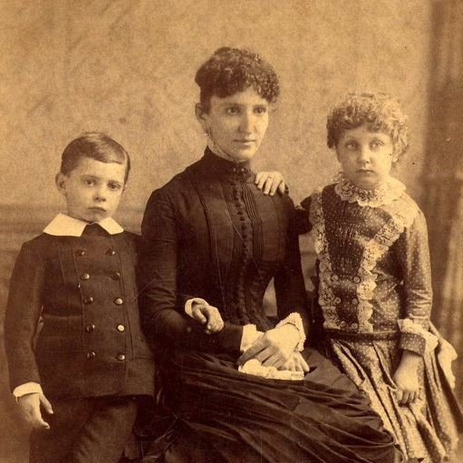 Image of young boy and girl standing next to their mother who is seated.