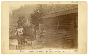 Sepia tone photograph of a woman standing outside of a wooden house. A wooded mountain is in the background.